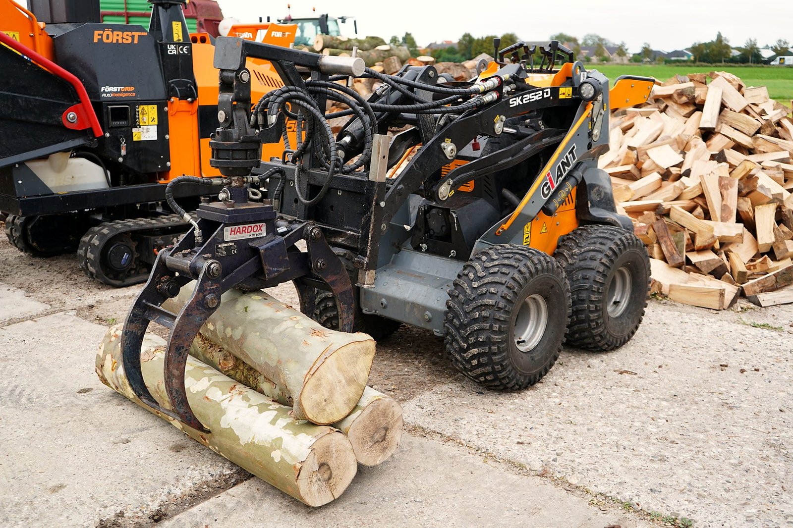 Minishovel met bomenklem tilt stammen op, met de rupsversnipperaar en haardhout op de achtergrond
